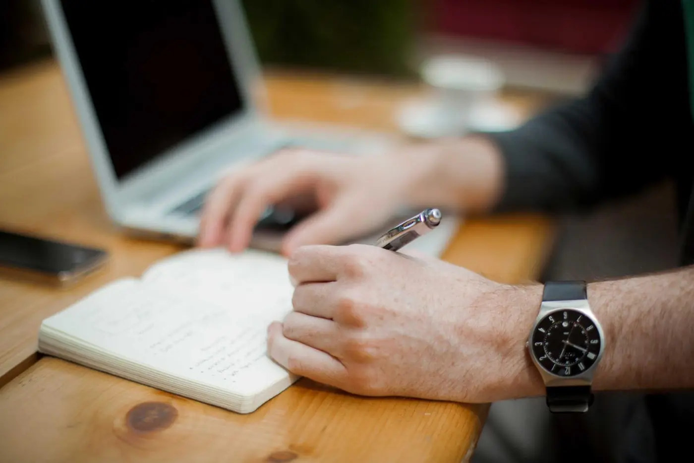 Person writing notes beside an open laptop